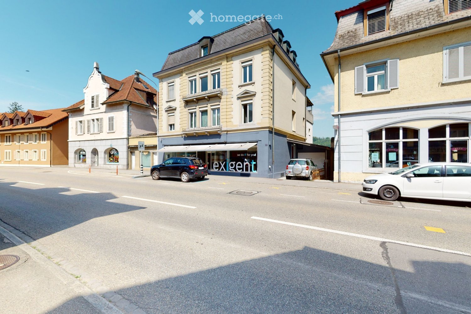 A street view showing a multi-story building with a storefront labeled "exgent" and several parked cars along the road.