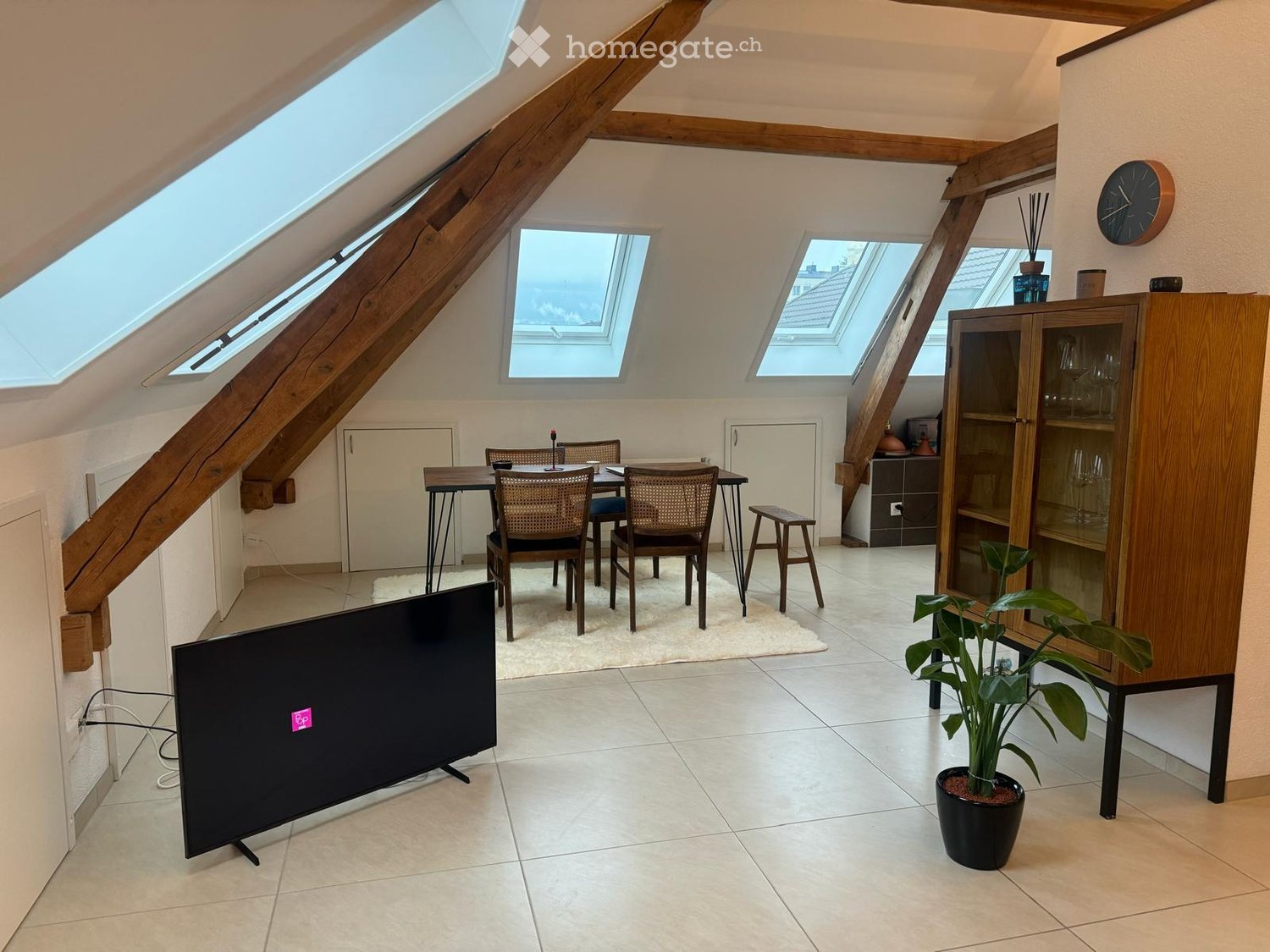 Bright attic room with skylights, tiled floor, television, dining table with chairs, cabinet, and potted plant.