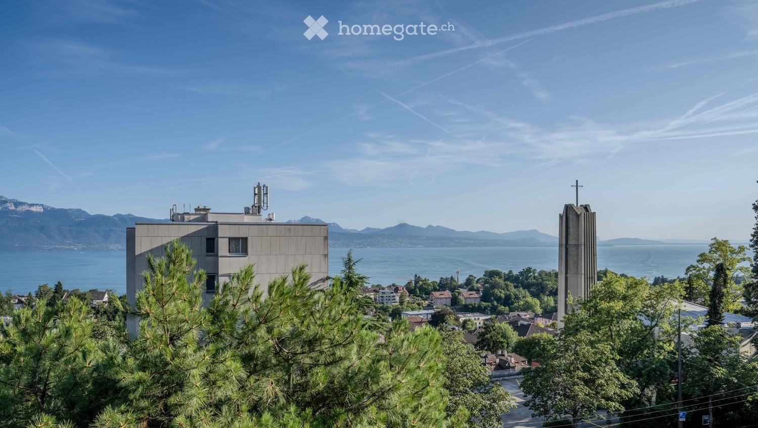 A modern building with a flat roof and several windows, surrounded by trees and overlooking a scenic lake and distant mountains.