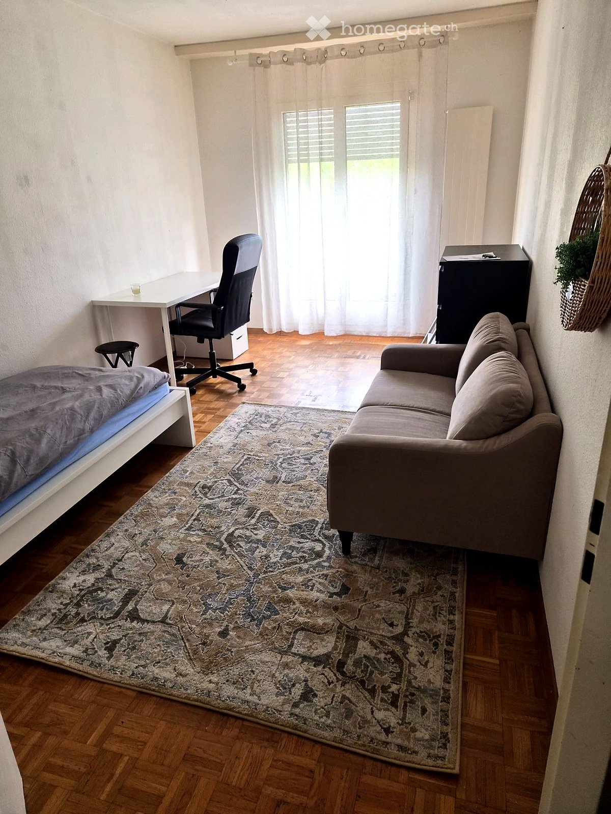 A compact, well-furnished room with a bed, sofa, desk, and chair. The room features wooden flooring, a patterned rug, and large windows with sheer curtains.