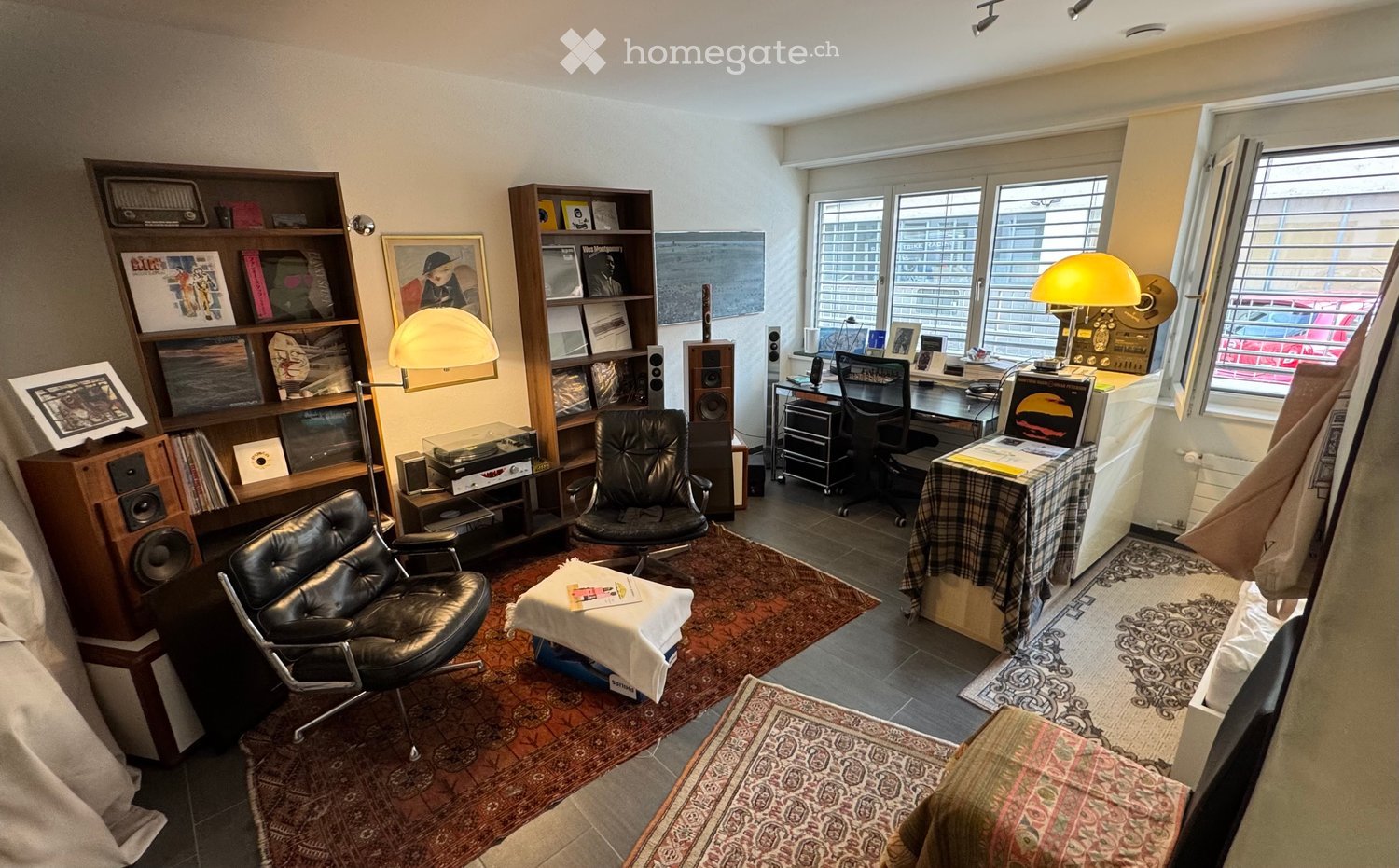 A well-furnished room with a large window, two black leather chairs, a small table with a white cloth, a bookshelf filled with books and records, a desk with a computer, and a lamp. The room features multiple rugs and a radiator.