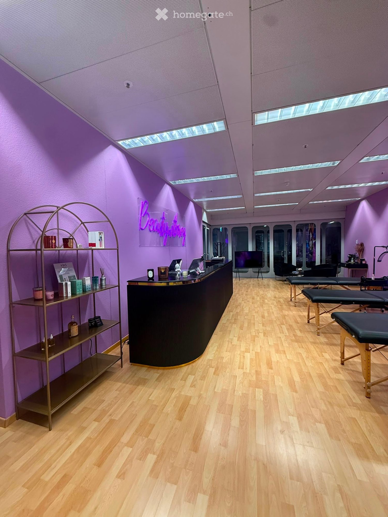 A modern beauty salon with purple walls, wooden flooring, a reception counter, shelves with products, and several massage chairs.
