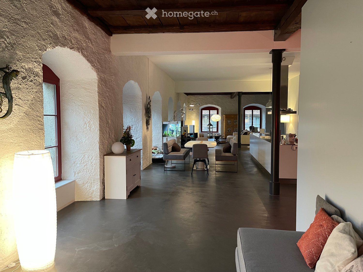 Modern, open-plan living area with exposed wooden beams, dark polished concrete floors, and a mix of contemporary and rustic furniture including a sofa, chairs, a table, and a tall floor lamp.