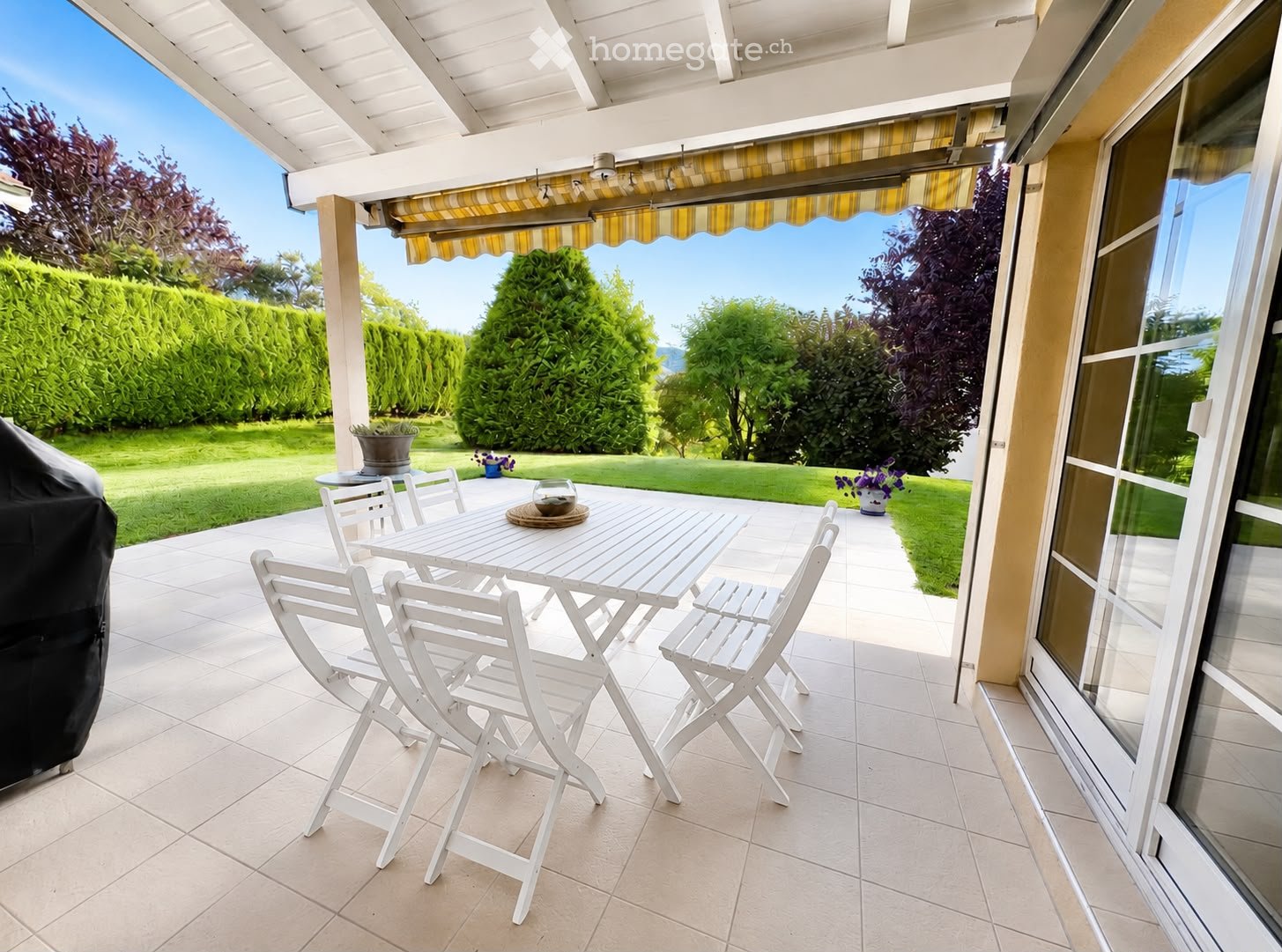 A covered patio with a white table and chairs, surrounded by a well-maintained garden and trees.