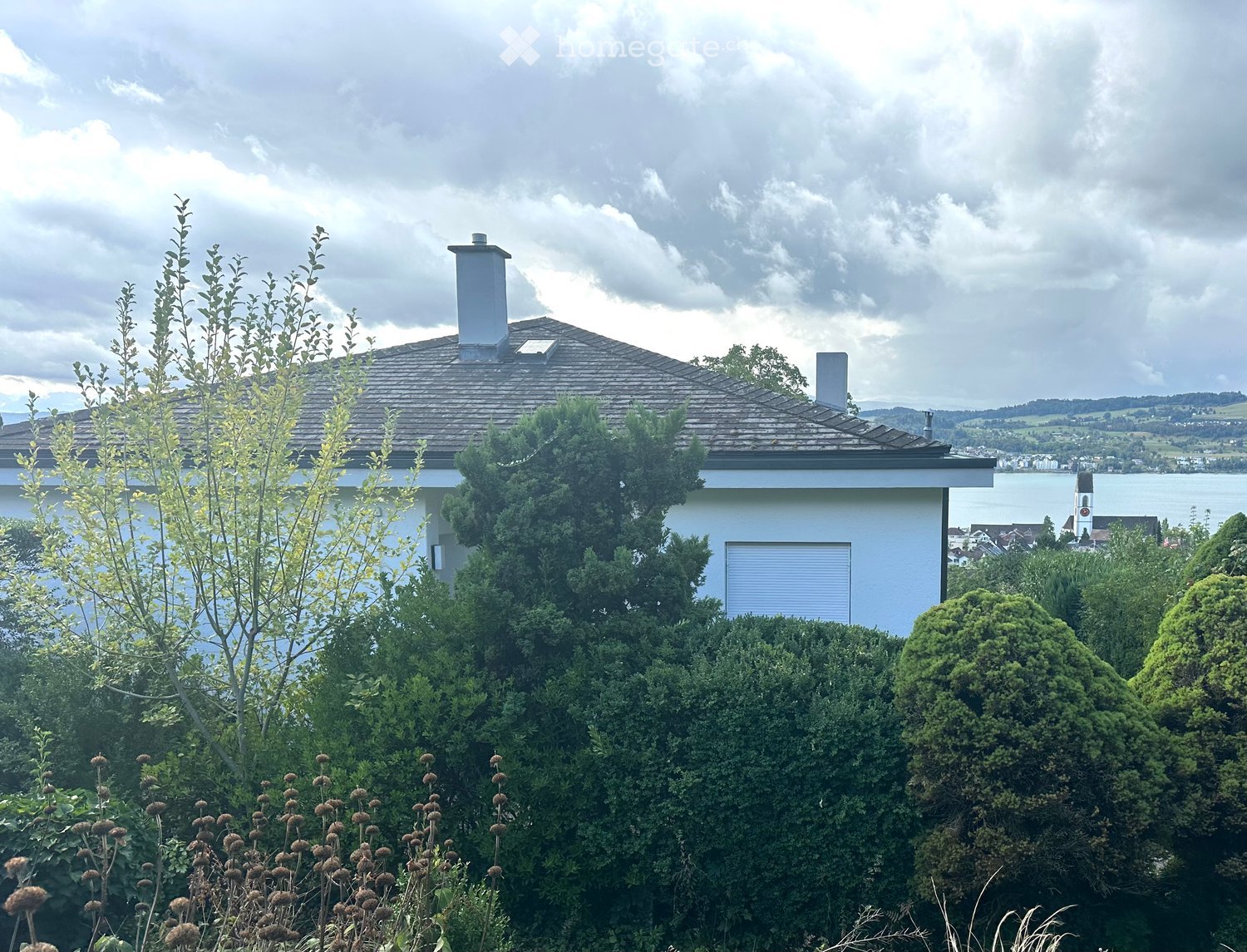A modern two-story house with a grey roof and white walls, surrounded by lush greenery and trees, with a scenic view of a lake and distant hills in the background.