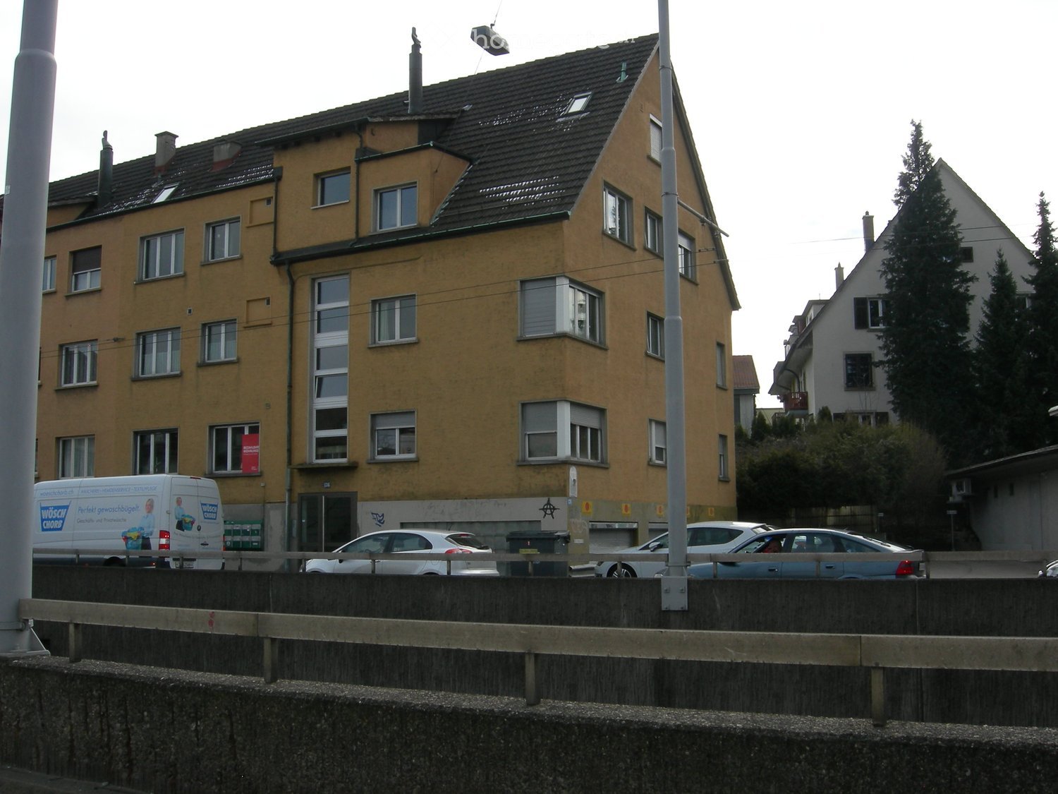 A multi-story residential building with yellow facade and multiple windows, located in an urban area with parked cars and a van nearby.