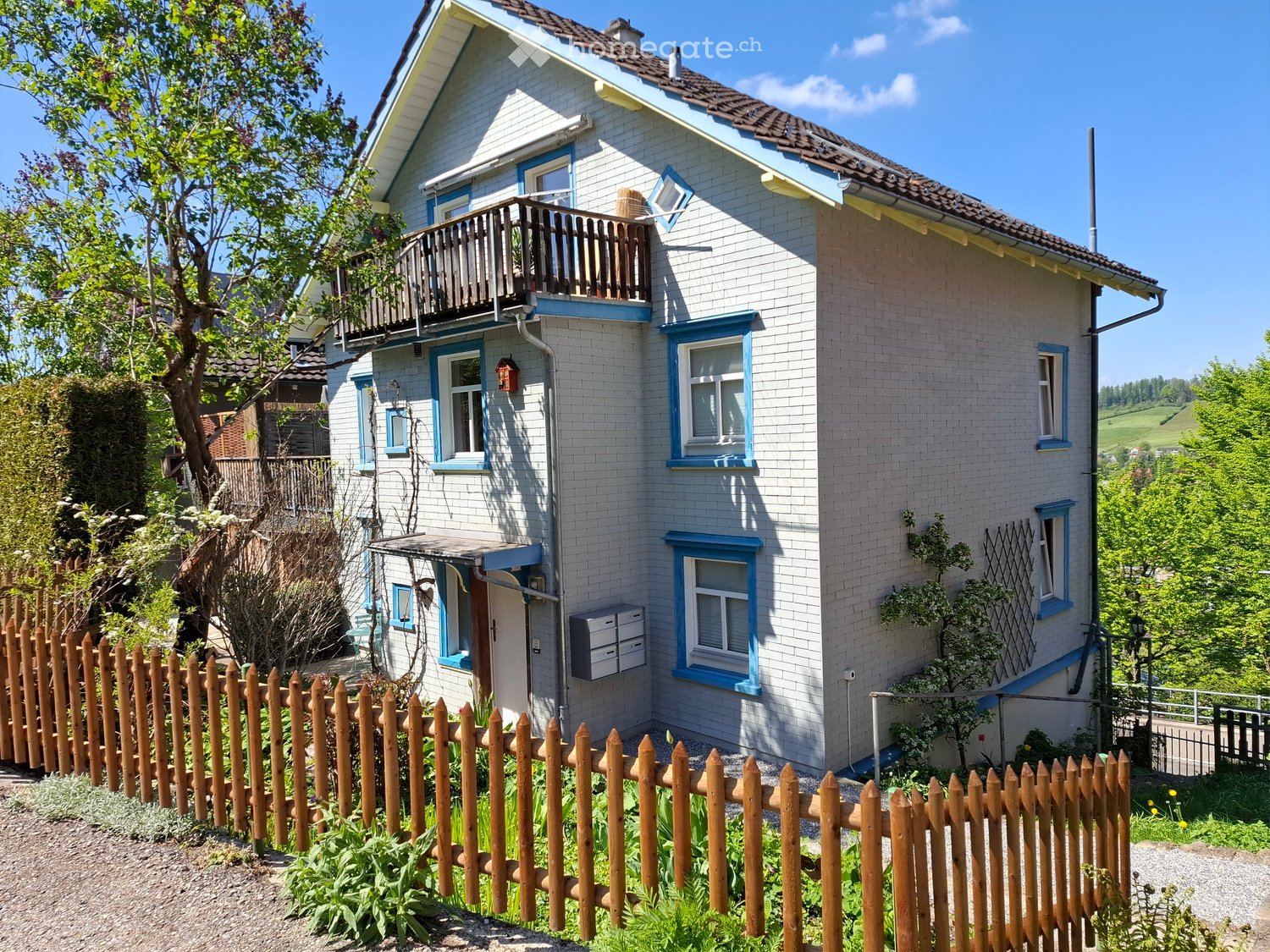 Two-story house with light blue siding, white trim, and multiple windows. Features a wooden balcony on the upper level and a small garden area enclosed by a wooden fence.