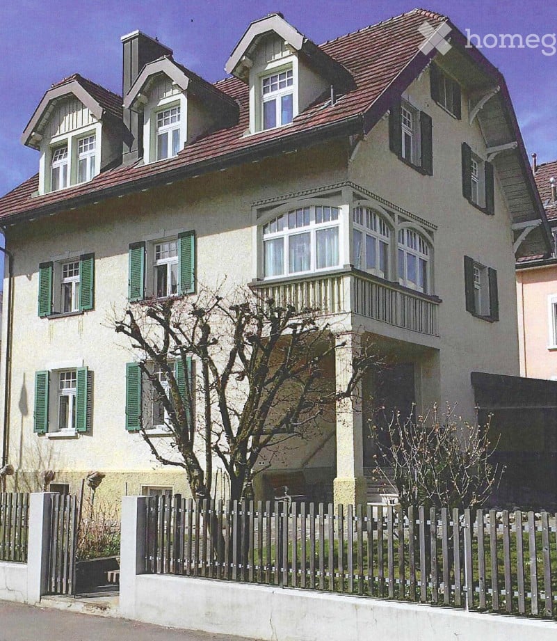 Two-story house with a red-tiled roof, multiple windows with green shutters, and a small balcony on the upper floor.
