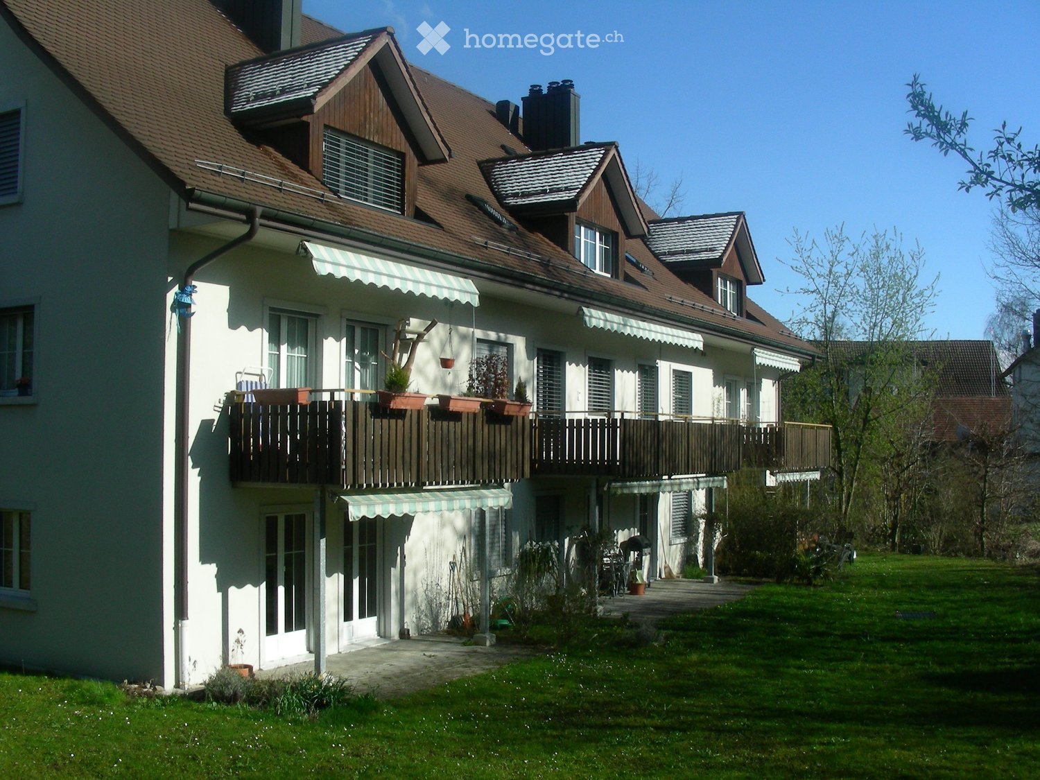 Two-story house with a brown tiled roof, white walls, multiple windows, and a balcony with potted plants. The house features a well-maintained lawn and garden area.