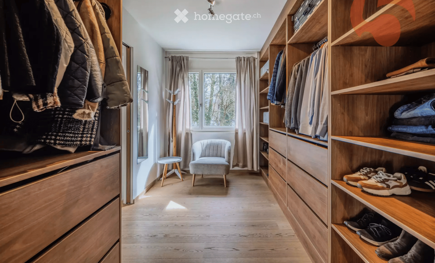 A well-organized walk-in closet with wooden shelves and drawers, a small seating area with a grey armchair and a round stool, and a coat rack near the window.