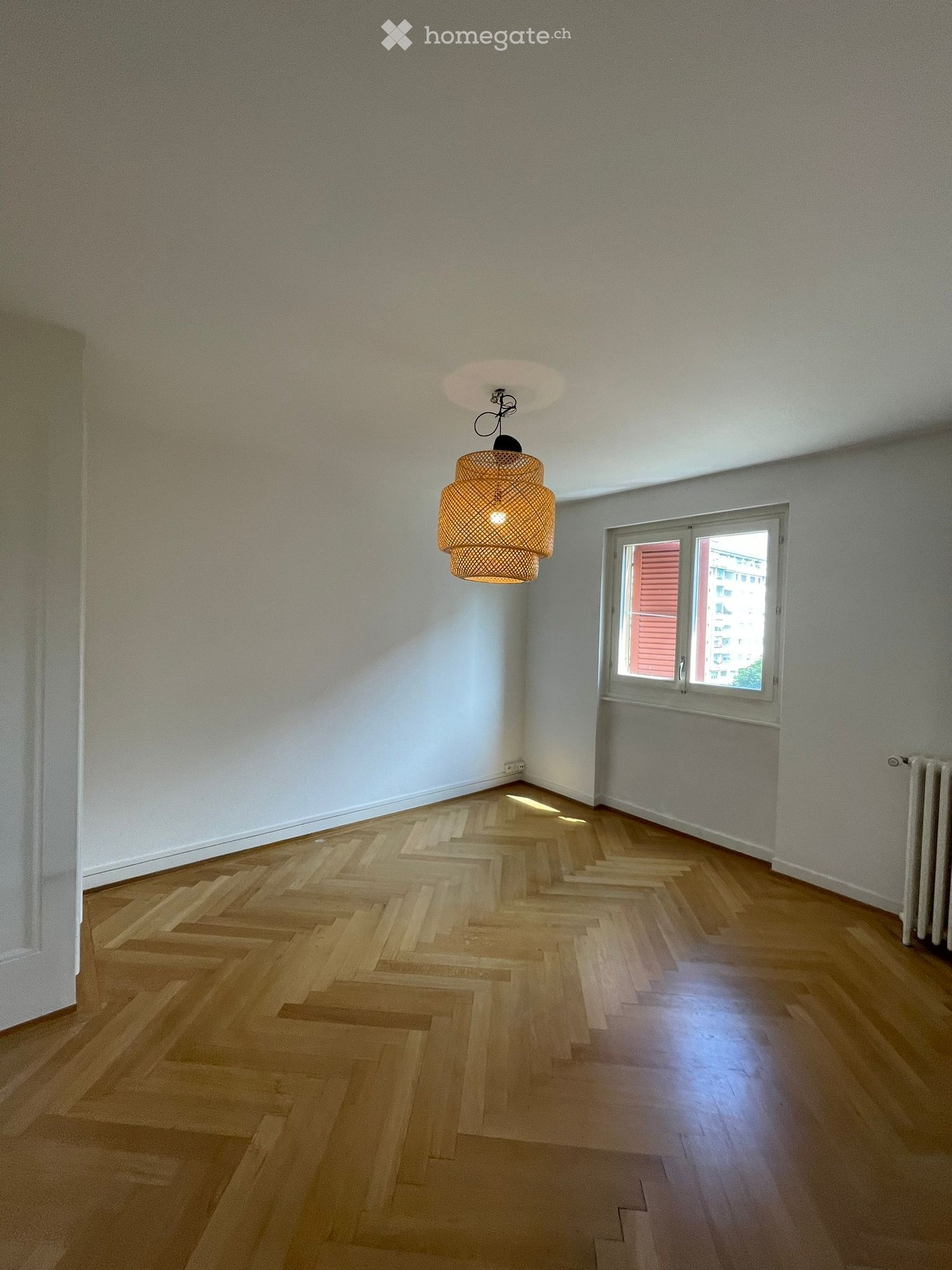 Empty room with wooden herringbone flooring, white walls, and a single hanging light fixture. A window with blinds offers natural light.