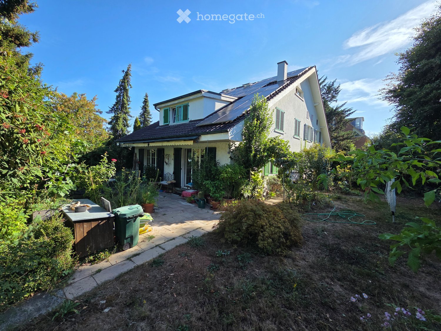 Garden area with a house in the background, featuring a patio, potted plants, a small table, and a trash bin.