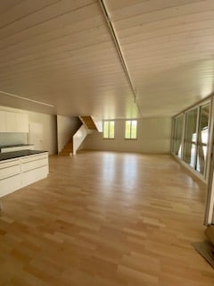 Bright, open-plan room with wooden flooring, white walls, and large windows. The space is completely empty, with no furniture or equipment visible.
