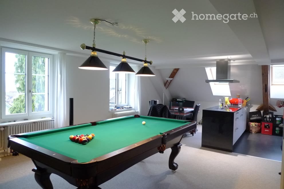 A spacious room with a pool table, surrounded by large windows and modern lighting. A kitchen area is visible in the background with cabinets and a counter.