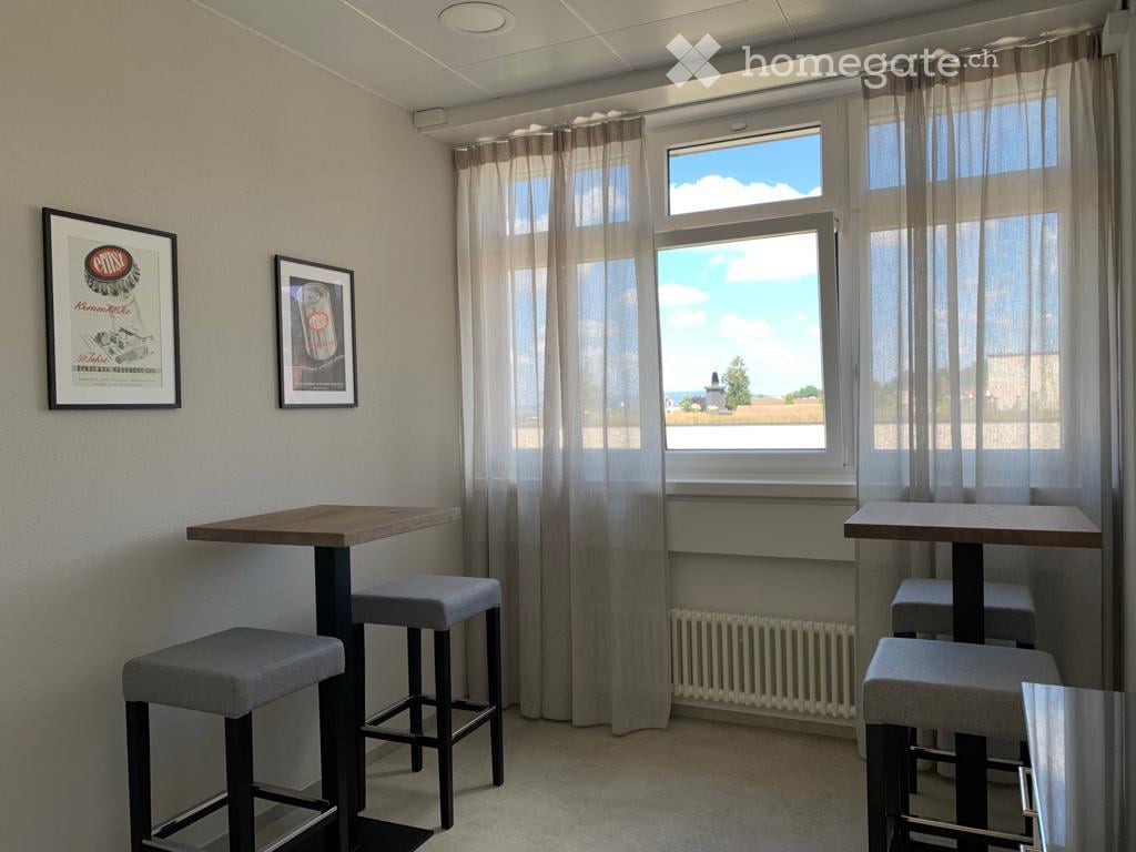 Bright room with two small tables and four stools, large windows with sheer curtains, and two framed pictures on the wall.