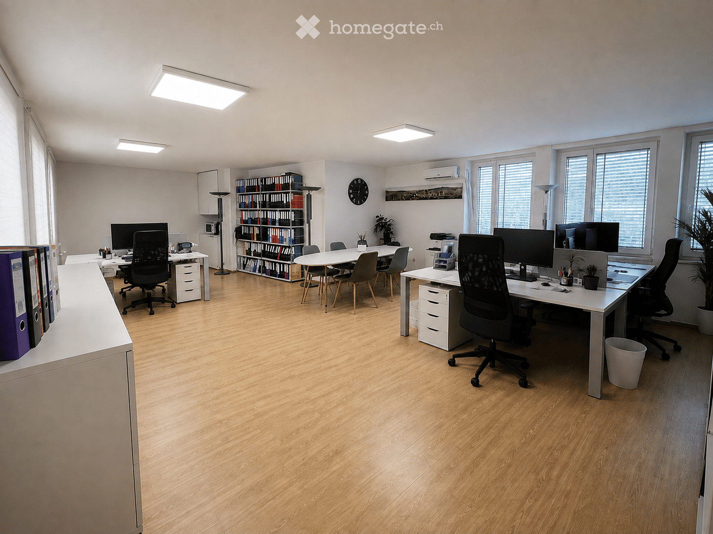 Modern office space with wooden flooring, multiple desks, chairs, computer screens, a bookcase, a clock, and potted plants.