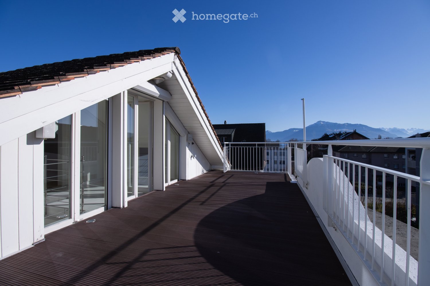 A modern rooftop terrace with wooden decking, white railings, and large glass windows leading to the interior. The background shows a clear blue sky and distant mountains.