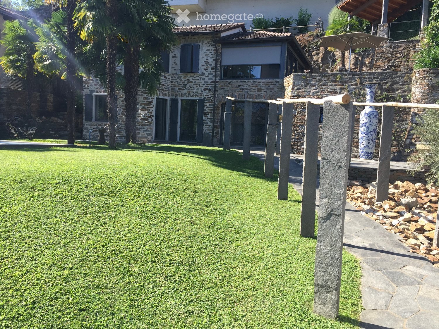 Well-maintained lawn with stone pillars, a decorative vase, and a stone pathway leading to a covered outdoor area with an umbrella. The house in the background is made of stone with large windows and a tiled roof.
