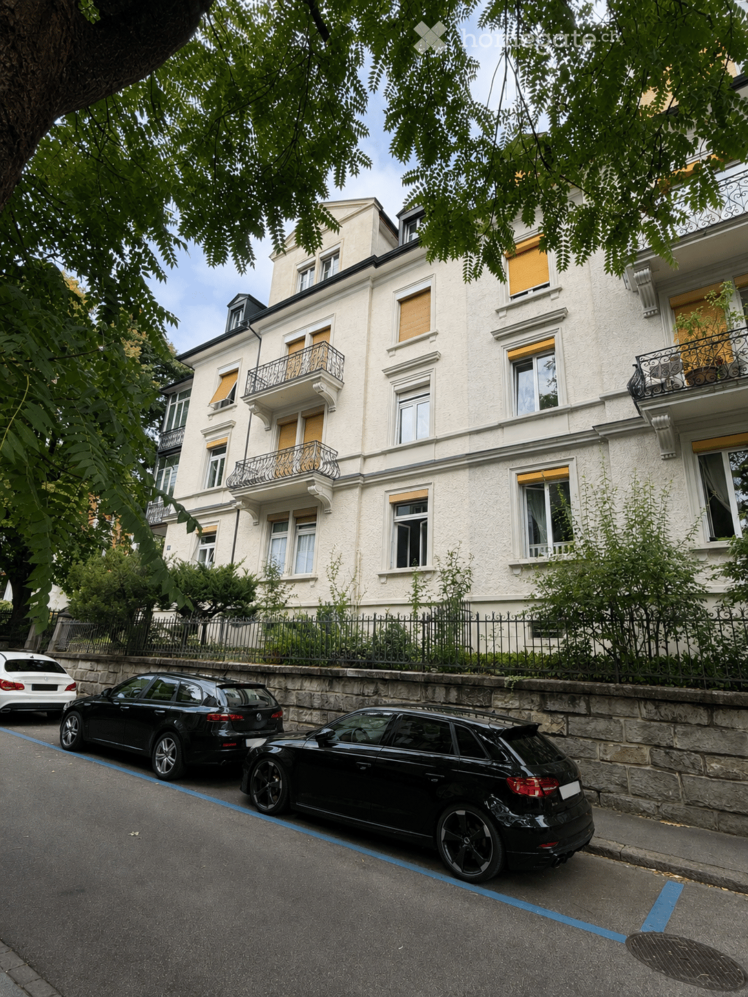A multi-story residential building with white facade, multiple windows, and balconies. The building is surrounded by greenery and has a stone fence along the sidewalk. Several cars are parked on the street in front of the building.