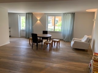 Bright living room with wooden flooring, large window with grey curtains, white sofa, dining table with chairs, and two small stools.