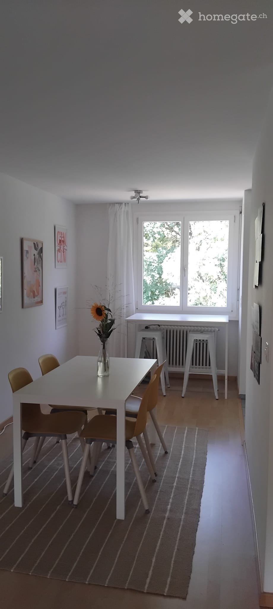 Dining area with a white table, four chairs, a vase with a sunflower, a white stool, and a radiator under the window.