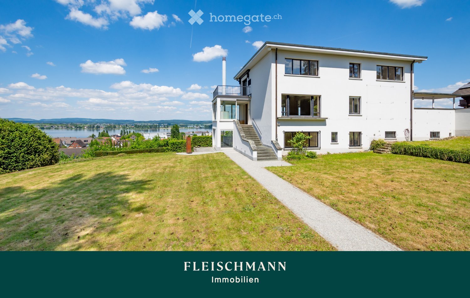 Modern two-story white house with large windows, a balcony, and a well-maintained lawn in front. The house features a staircase leading to the entrance and is situated near a body of water with distant hills.
