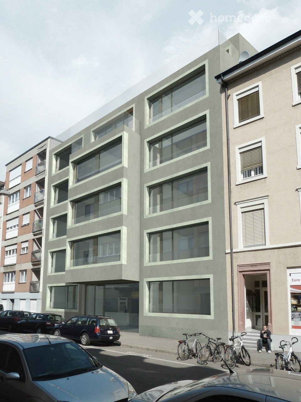 Modern multi-story building with large windows and balconies, located on a street with parked cars and bicycles.