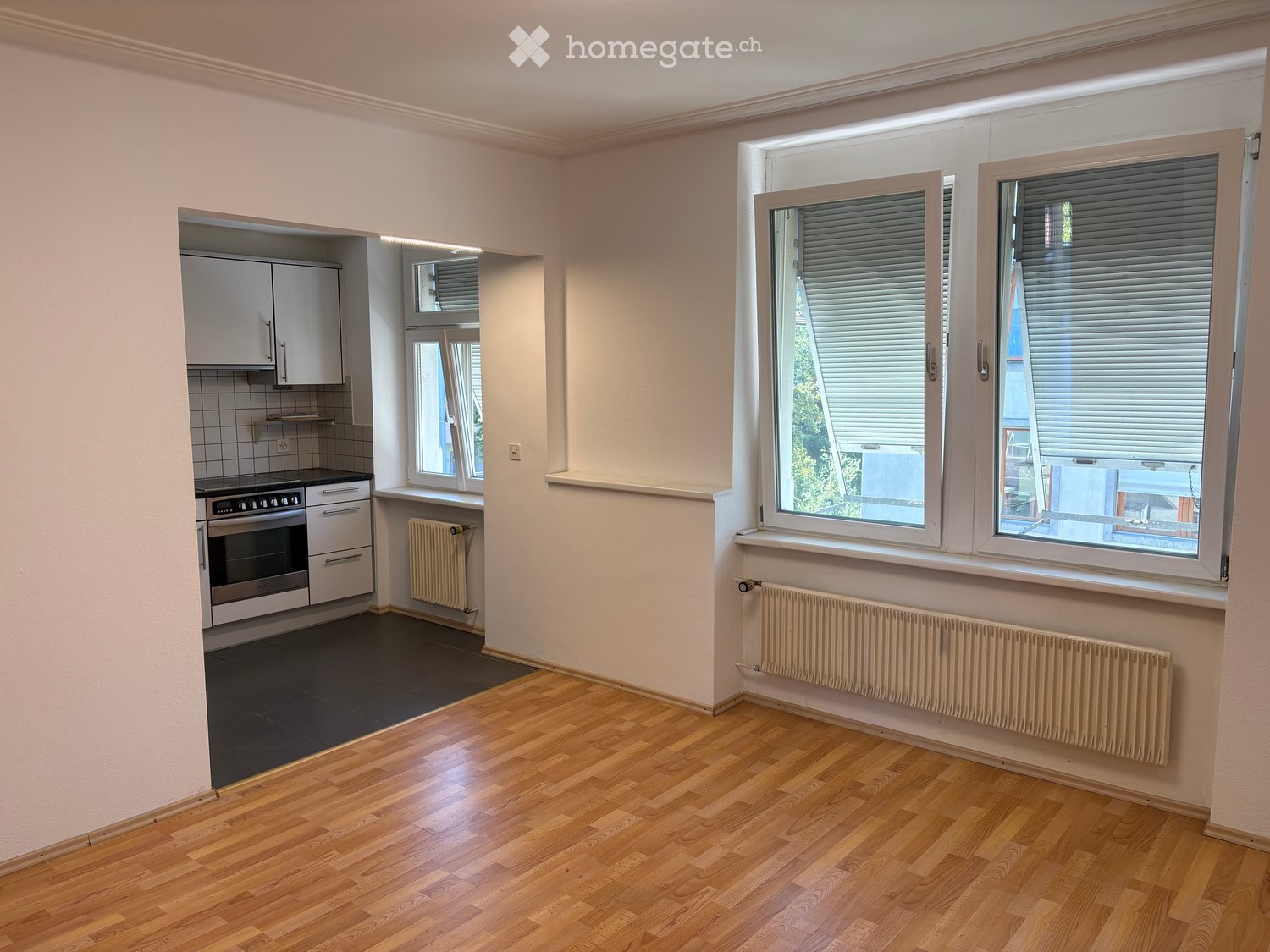 Bright living space with wooden flooring, large windows, integrated kitchen area with oven and cabinets, and radiators along the walls.