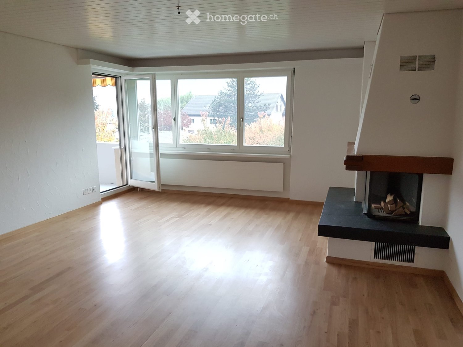 Empty room with wooden flooring, large windows, and a fireplace with wood logs inside.