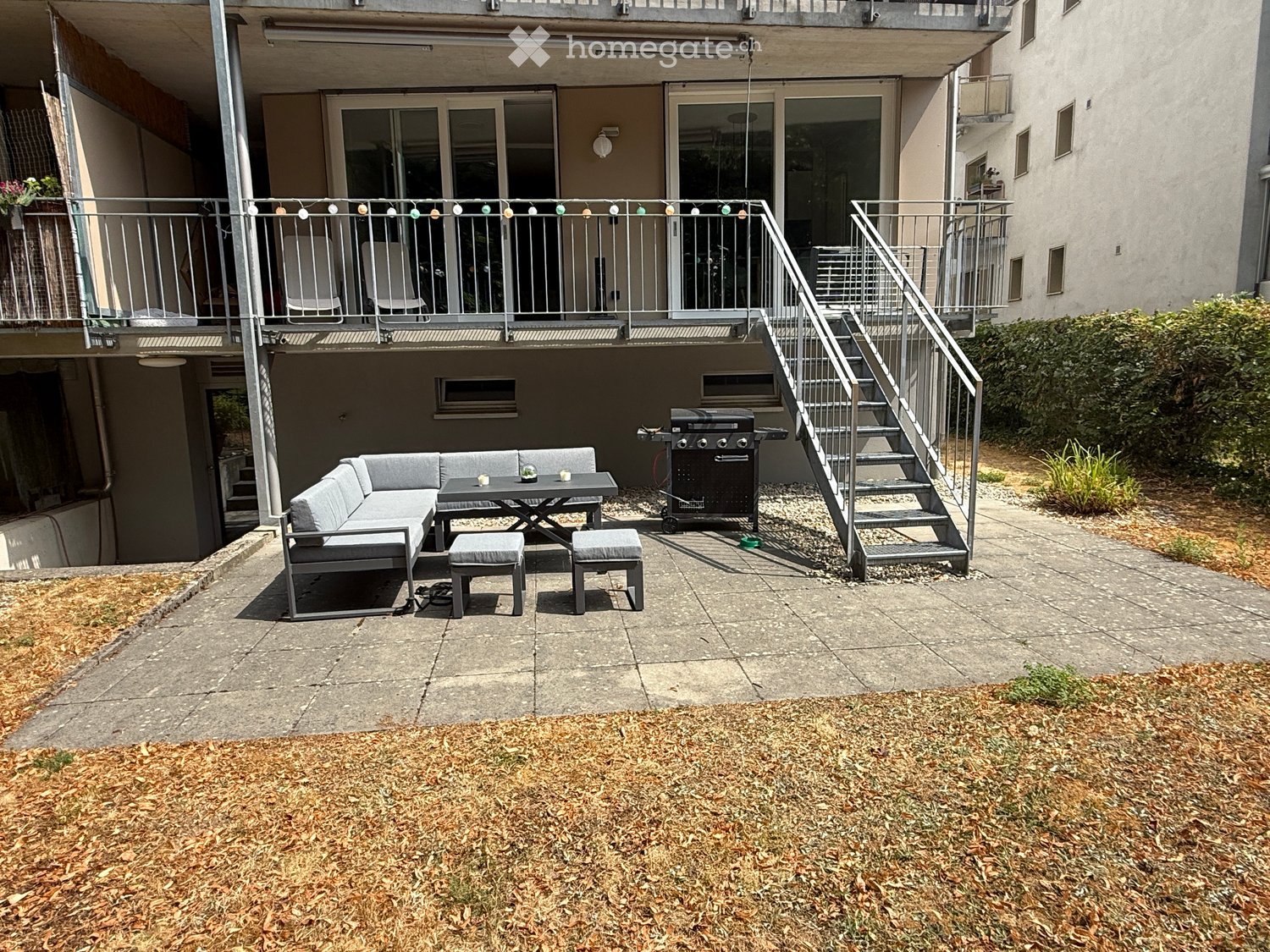 Outdoor patio with a grey sectional sofa, small table, two stools, and a grill. Metal staircase leads to an upper balcony with chairs and a railing.