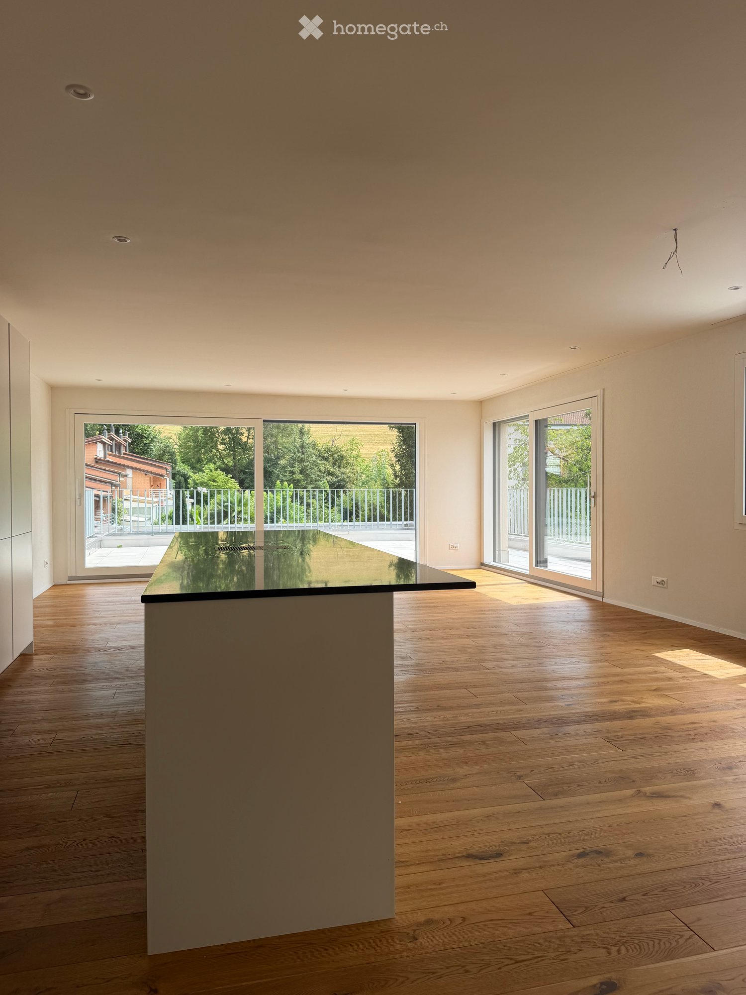 Empty room with wooden flooring, large glass doors leading to a balcony, and a central island with a black countertop.