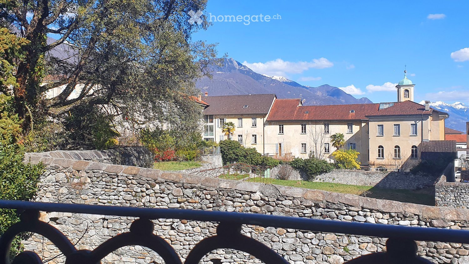 A scenic view of a town with traditional buildings, red-tiled roofs, and a stone wall in the foreground, with mountains in the background.