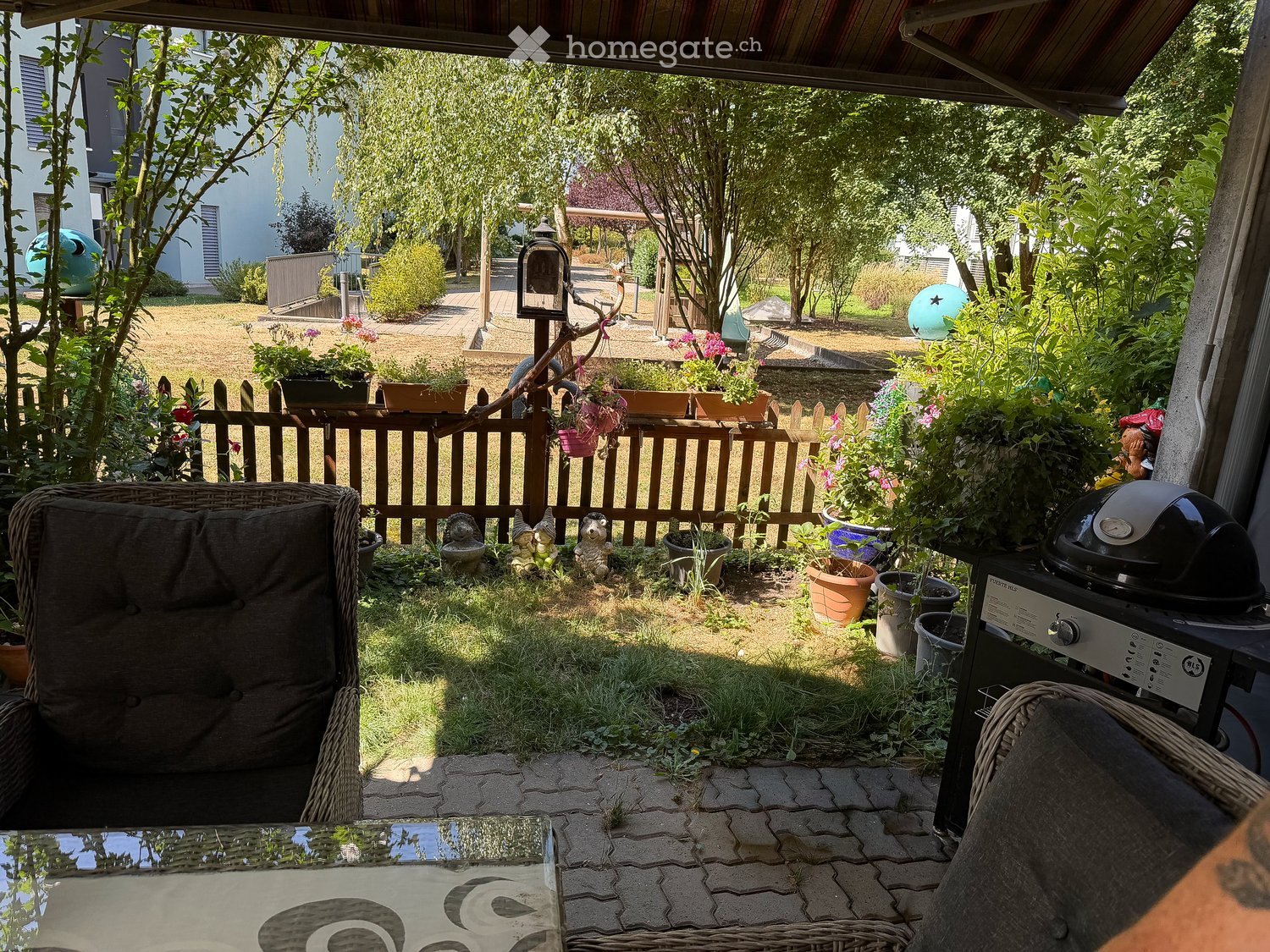 Garden area with a wooden fence, potted plants, garden ornaments, a barbecue, and a covered seating area with wicker chairs and a glass table.