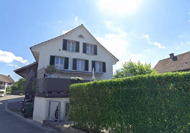 Two-story white house with black shutters, multiple windows, and a small balcony. The house has a well-maintained garden with a hedge in the front and a small staircase leading to the entrance.