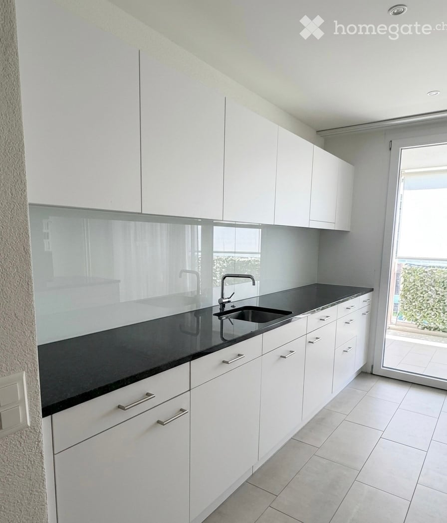 Modern kitchen with white cabinets, black countertop, and a sink with a modern faucet. The space is bright with large windows and a sliding glass door leading to a balcony.