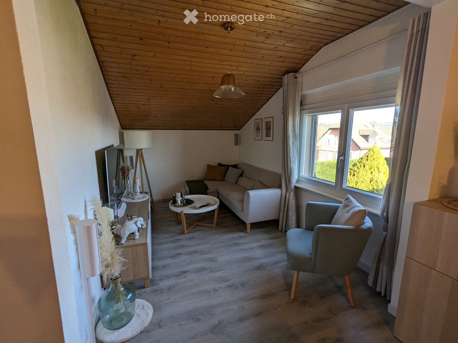 Modern living room with wooden floors, white walls, and large windows. Features a white sofa, coffee table, armchair, and decorative items including a vase and picture frames.