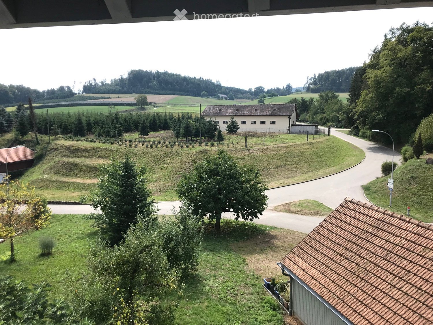View from a balcony or terrace showing a landscaped garden area with trees, a pathway, and a distant building in a rural setting.