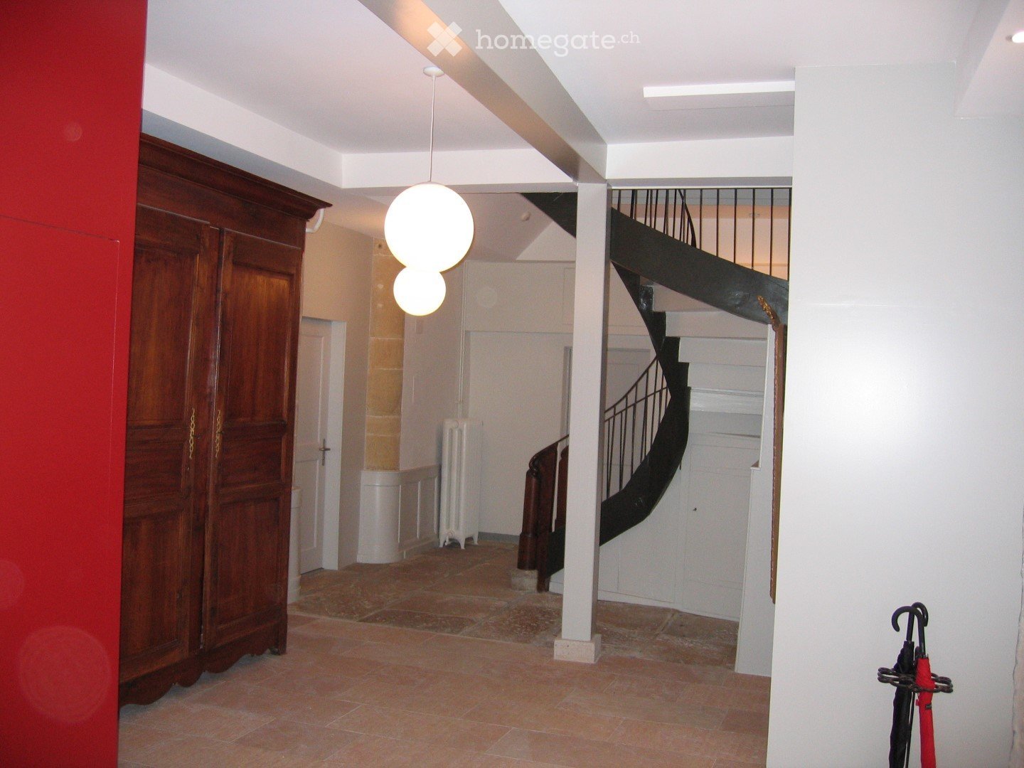 Empty hallway with tiled floor, white walls, a wooden wardrobe, a staircase with black railing, and a hanging light fixture.