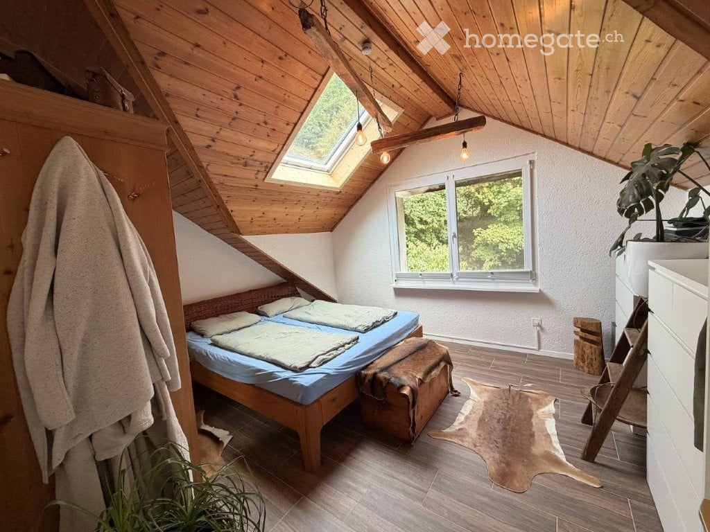 Cozy attic bedroom with wooden slanted ceilings, skylight, large window, double bed, wardrobe, and a small plant.