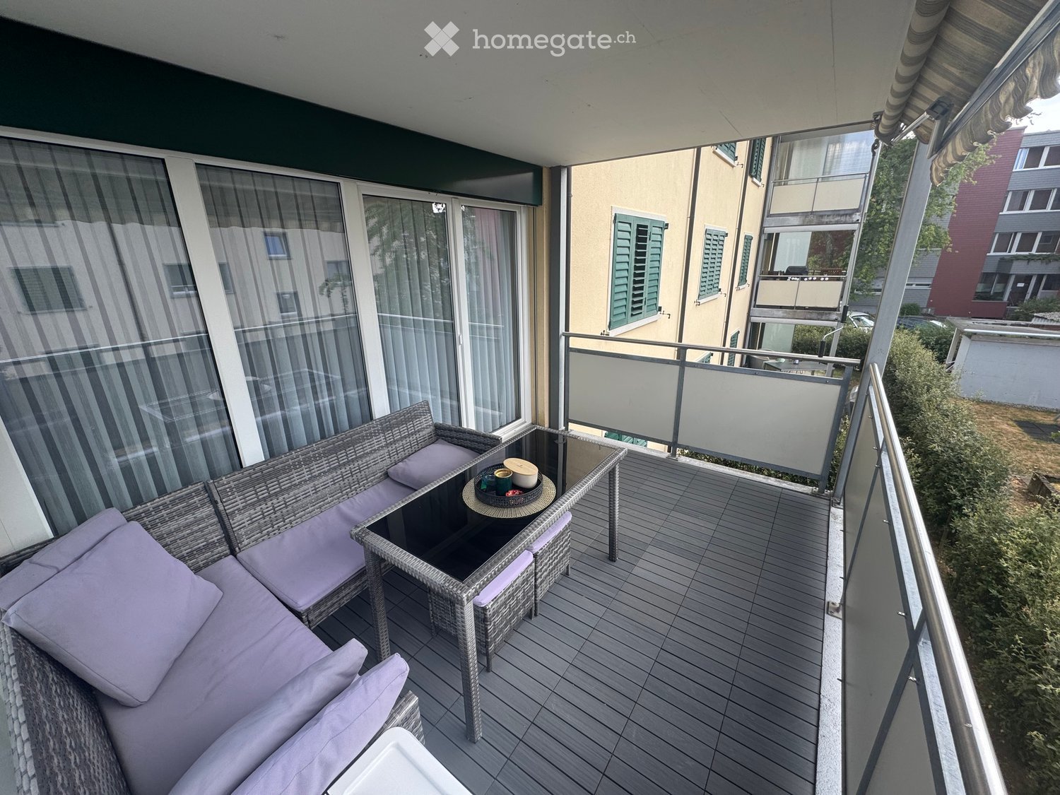 Balcony with wooden decking, wicker sofa with cushions, glass table, and small round tray with cups.