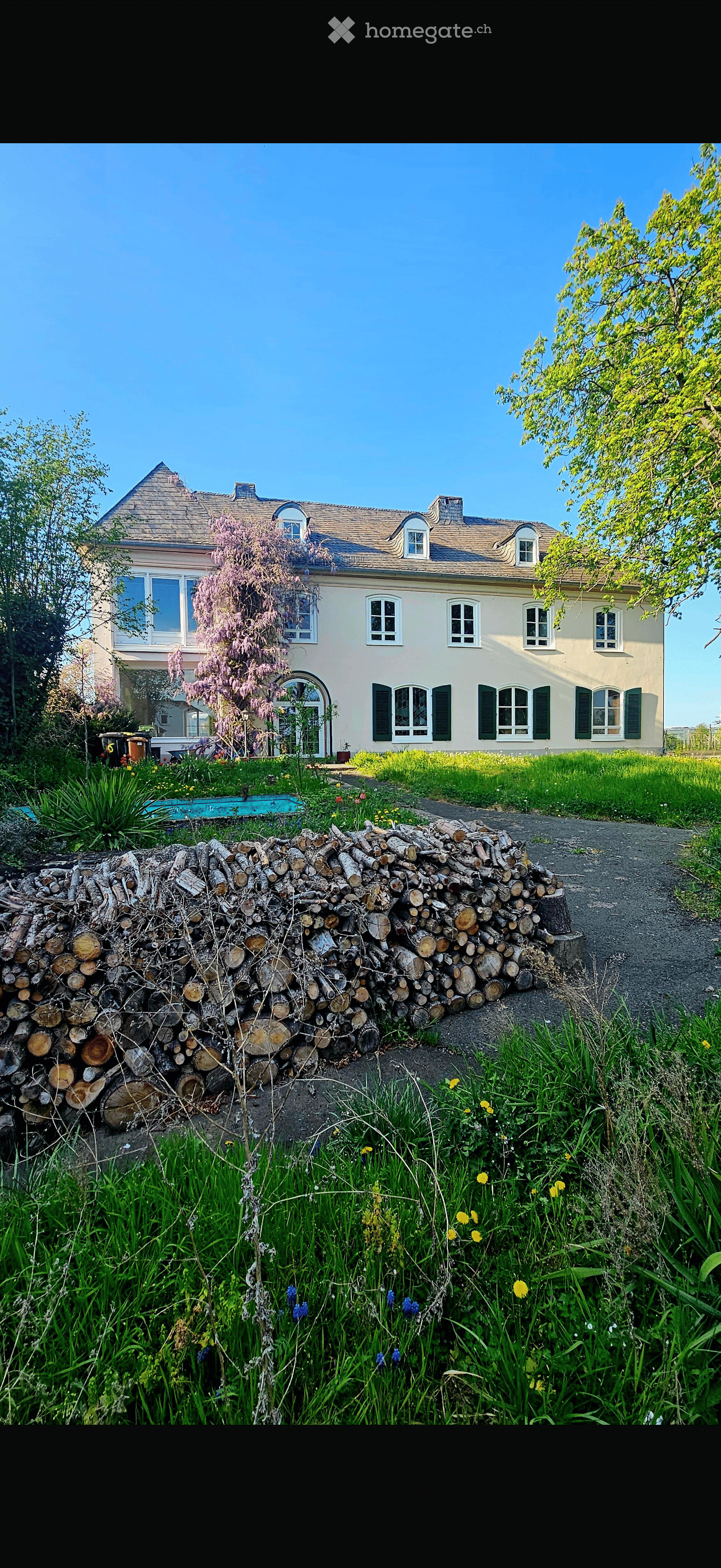 A large, well-maintained garden with a stack of firewood, a swimming pool, and a two-story house with multiple windows and a sloped roof.