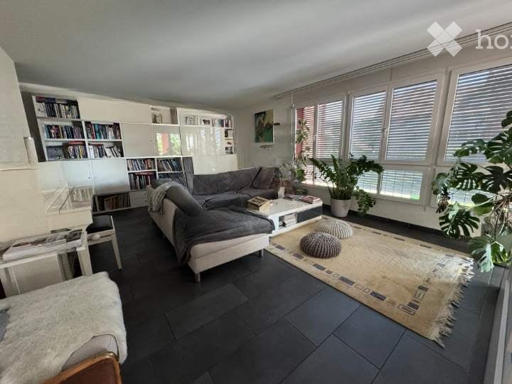 Modern living room with large windows offering a view, featuring a grey sectional sofa, white coffee table, ottomans, bookshelf, and plants.