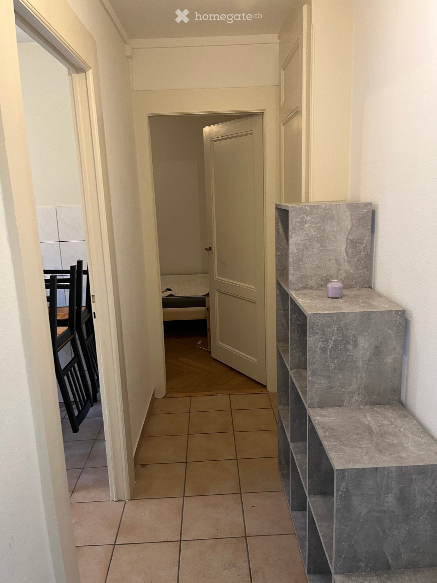 A narrow hallway with tiled flooring, white walls, and a marble bench on the right side. A door leads to another room, and a glimpse of a dining area is visible on the left.