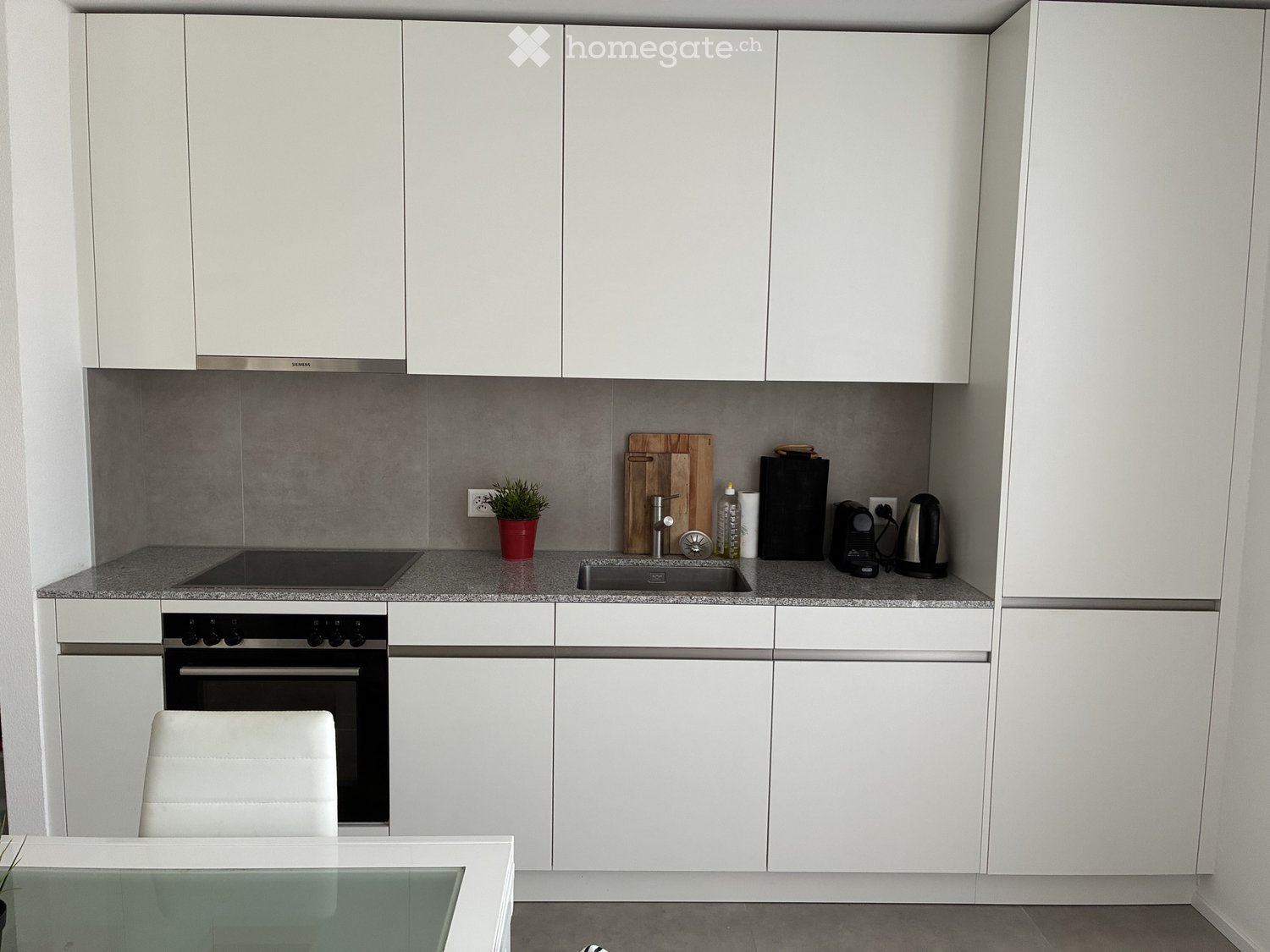 Modern kitchen with white cabinets, grey countertops, built-in oven, sink, and various appliances including a coffee maker and kettle. A small potted plant and a wooden cutting board are on the counter.