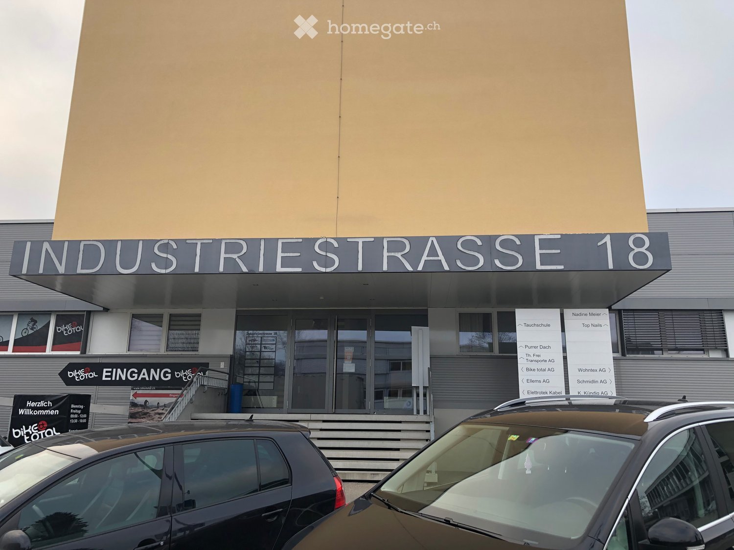 Modern commercial building with large glass windows, signage reading "INDUSTRIESTRASSE 18", and several parked cars in front.