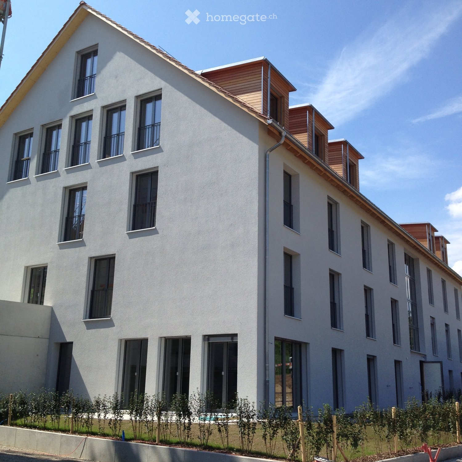 Modern two-story building with white facade, multiple windows, and balconies. The roof features wooden accents and small dormer windows.