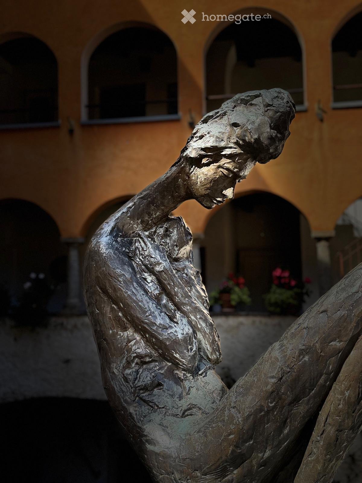Bronze sculpture of a human figure in a dynamic pose, set against a backdrop of a historic building with arched windows and potted plants.