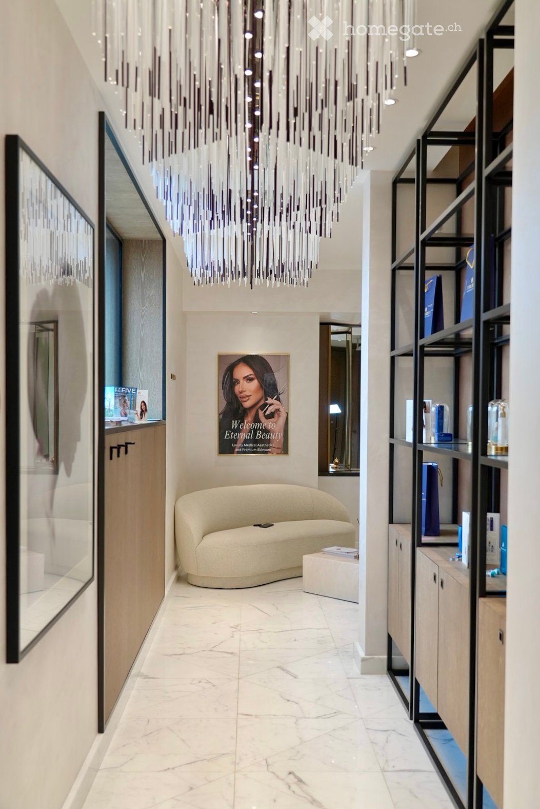 Modern, well-lit hallway with a large chandelier, a cream-colored sofa, a small table, shelves with products, and a welcome sign.