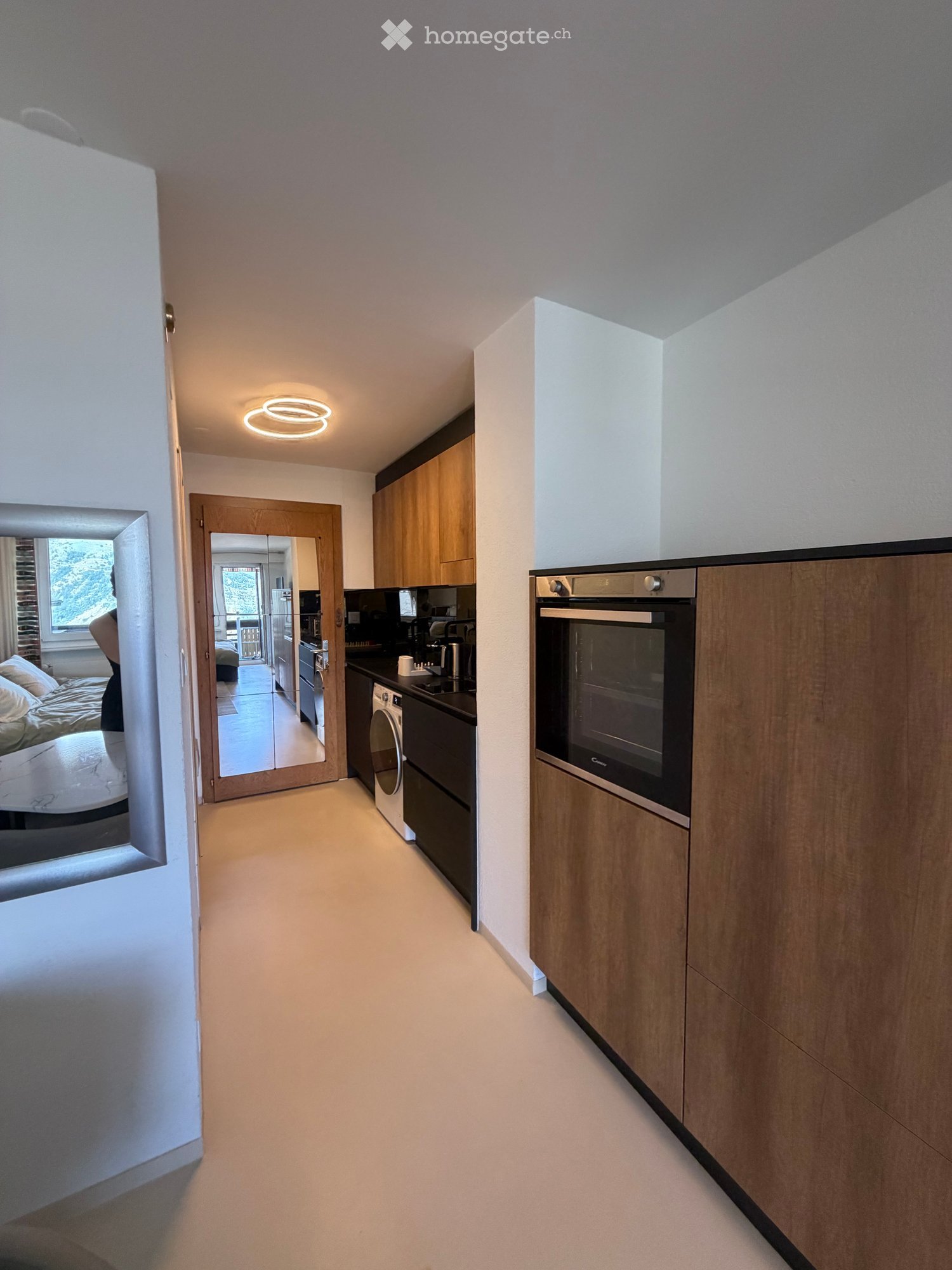 Modern hallway with light-colored walls, wooden cabinets, and a circular ceiling light. A mirror reflects a view of a bedroom and kitchen area.