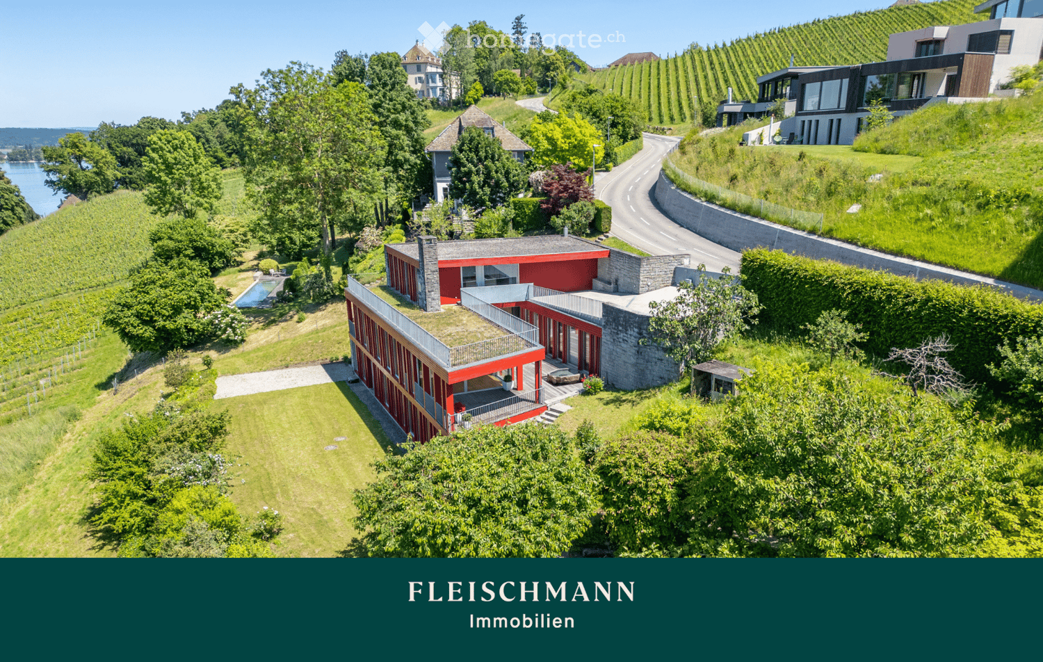 Aerial view of a modern red and grey house with a green roof, surrounded by lush greenery, vineyards, and a swimming pool. The house features large windows and a spacious terrace, with a scenic view of the surrounding landscape.